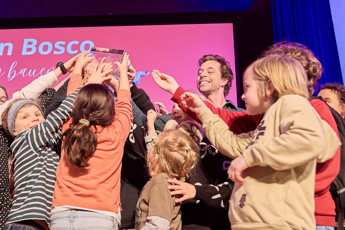 Lächelnder Mann und aufgeregte Kinder auf der Bühne, die gemeinsam eine durchsichtige Trophäe halten und einen Erfolg feiern.