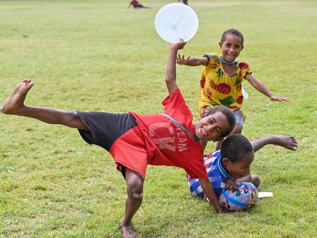 Drei fröhliche Kinder spielen auf einer Wiese. Eines balanciert auf einem Bein mit Frisbee, ein anderes lächelt breit.