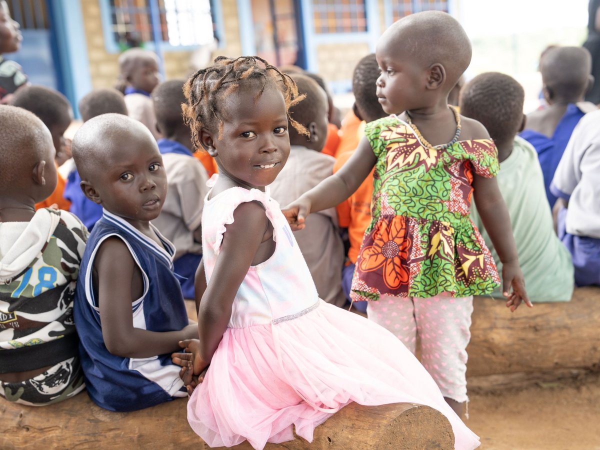 Drei junge afrikanische Kinder auf einem Baumstamm: ein lächelndes Mädchen in Rosa, ein Junge und ein Mädchen im gemusterten Kleid.