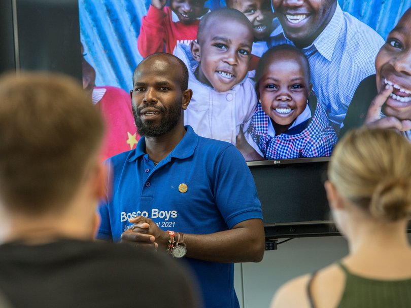 Ein Mann im 'Bosco Boys' Hemd spricht; dahinter ein Bildschirm mit lächelnden afrikanischen Kindern und einem Erwachsenen.