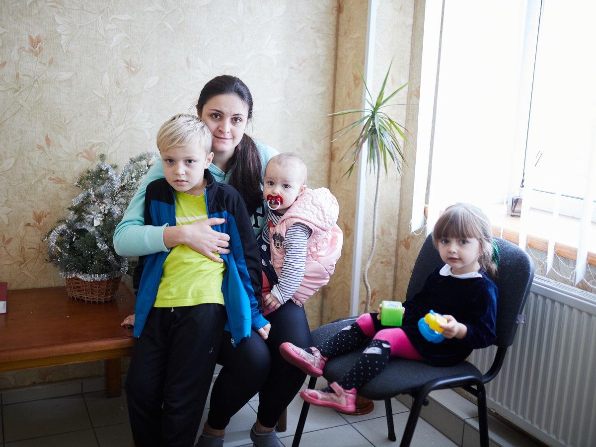 Eine Frau mit drei Kindern im Innenbereich, darunter ein Junge, ein Baby mit Schnuller und ein Mädchen mit Spielzeug.