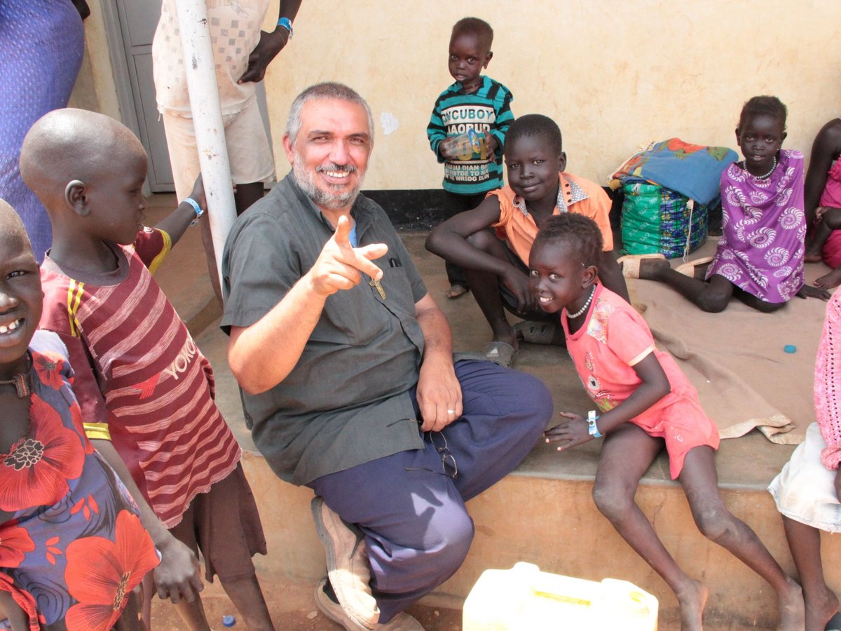 Lächelnder Mann zeigt auf Kamera, umgeben von afrikanischen Kindern in einfacher Umgebung.