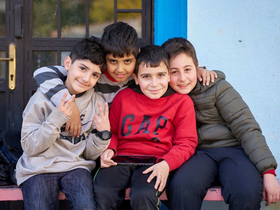 Vier fröhliche Jungen sitzen nebeneinander, umarmen sich, lachen in die Kamera. Einer hält ein Handy und zeigt ein Friedenszeichen.
