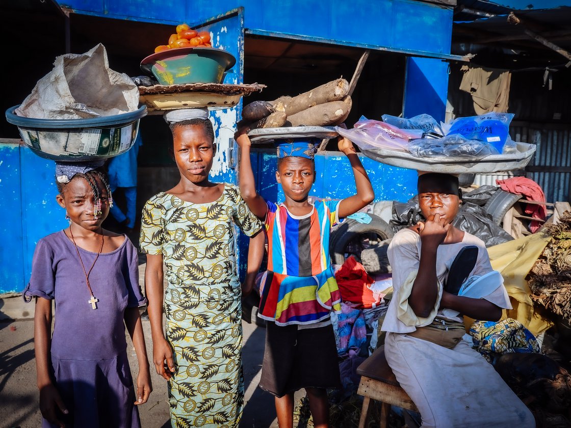 Vier junge afrikanische Kinder balancieren verschiedene Waren wie Tomaten, Yams und verpackte Artikel auf dem Kopf am Markt.
