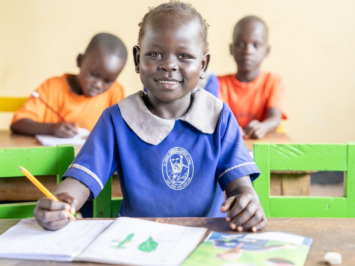 Lächelndes Schulkind in blauer Uniform, hält Bleistift, sitzt am Schreibtisch im Klassenzimmer, andere Kinder im Hintergrund.