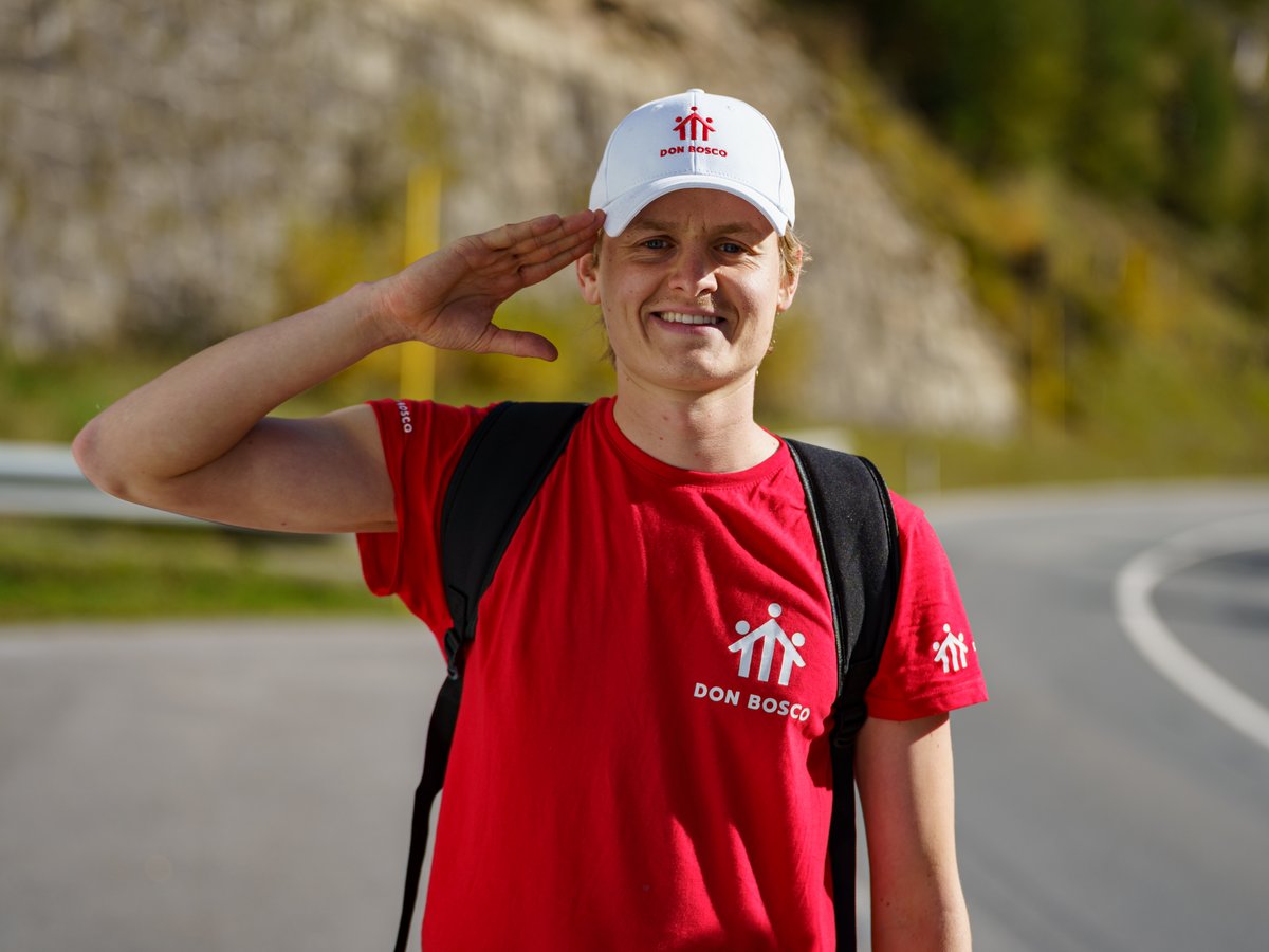 Ein lächelnder Mann in rotem Don Bosco T-Shirt und weißer Mütze salutiert, mit Rucksack und verschwommenem Straßen-/Hügelhintergrund.