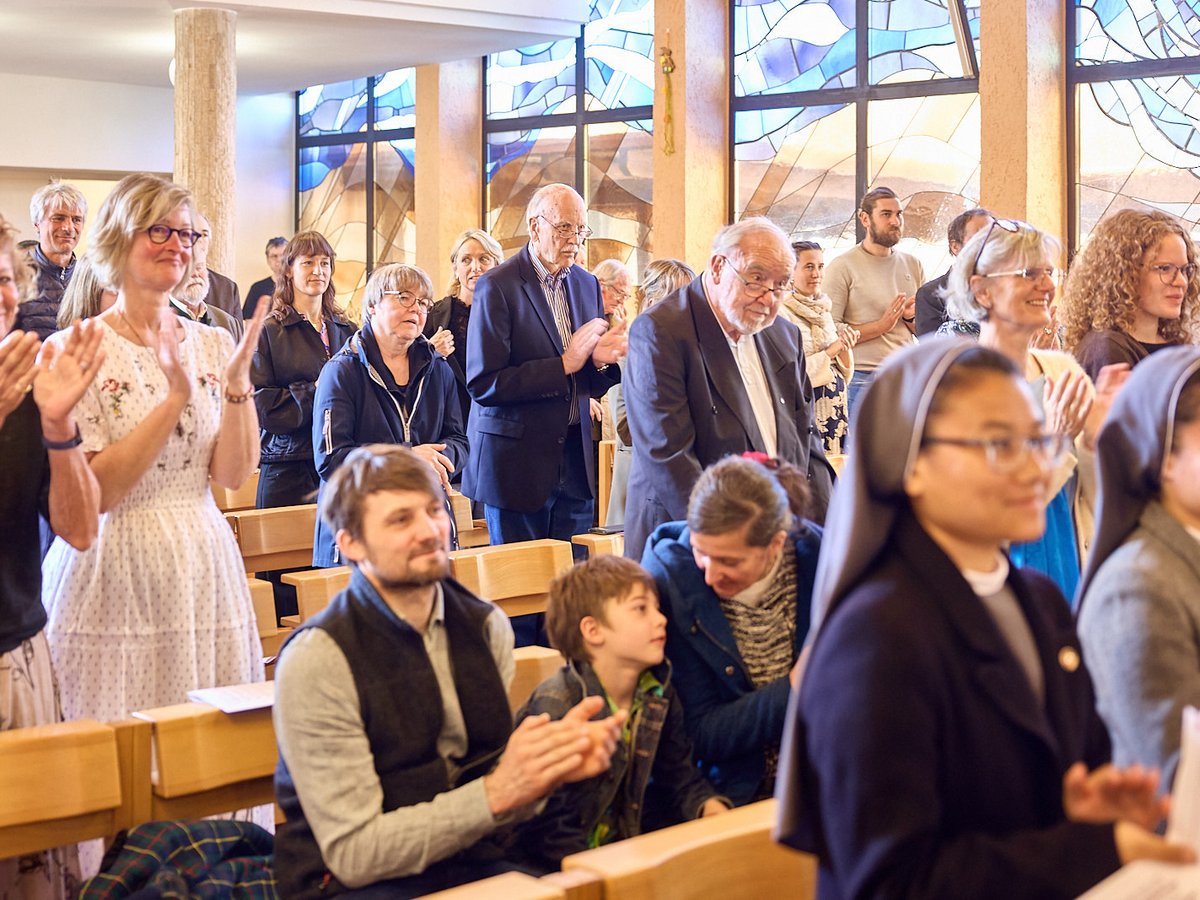 Eine diverse Gruppe von Menschen, darunter Nonnen und Familien, klatscht und lächelt in einer Kirche mit Buntglasfenstern.