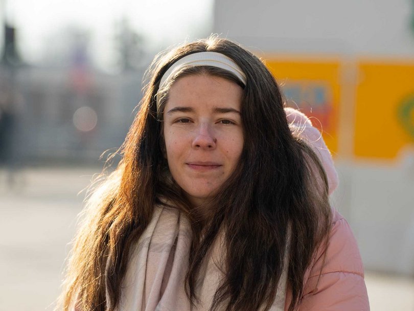 Junge Frau mit langen dunklen Haaren, weißem Stirnband, rosa Jacke und Schal blickt nach vorne im Freien.