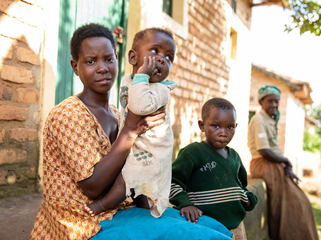Eine junge Frau hält ein Baby; ein weiteres Kind blickt in die Kamera. Szene in ländlicher afrikanischer Umgebung.