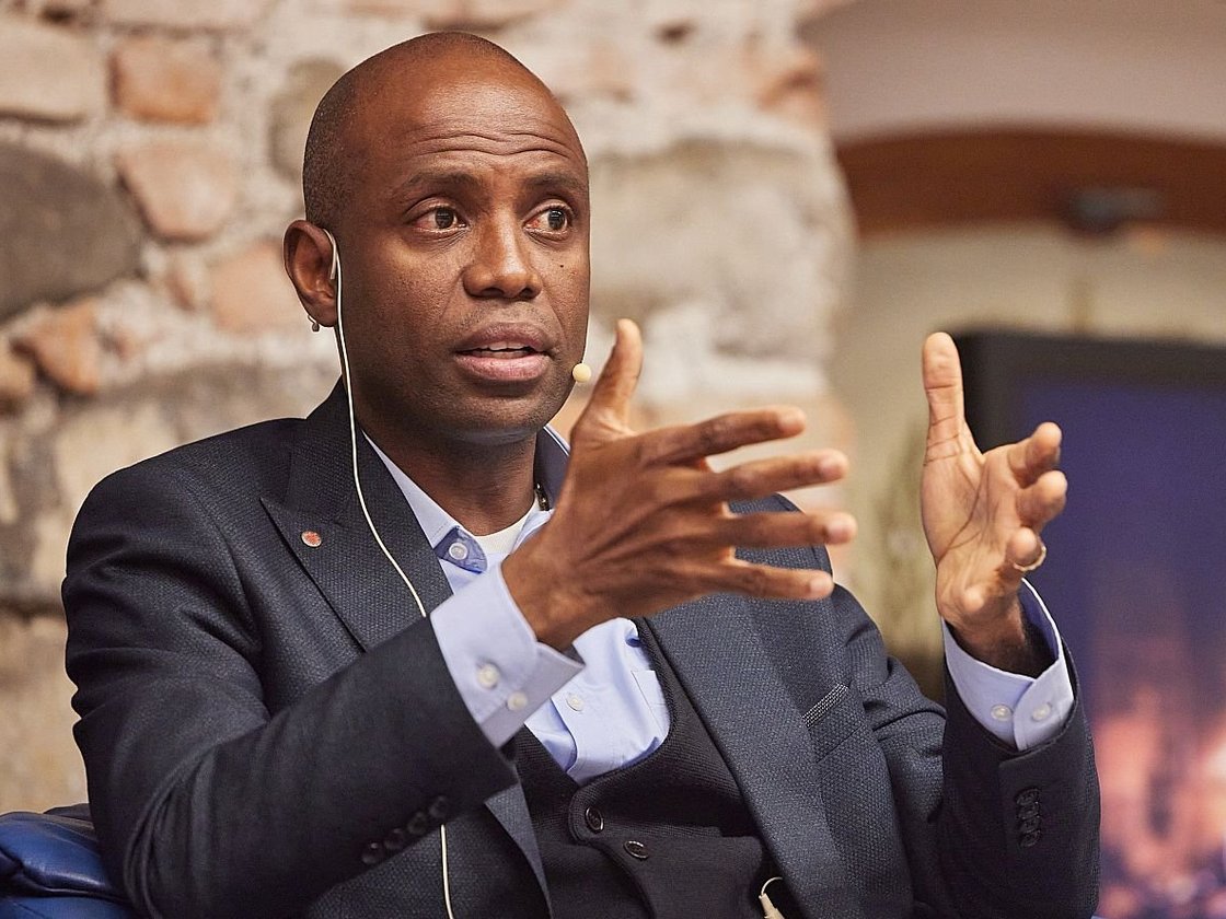 Ein Mann spricht lebhaft mit ausgestreckten Händen, trägt Anzug und Ohrhörer, vor einer Steinwand und unscharfem Bildschirm.