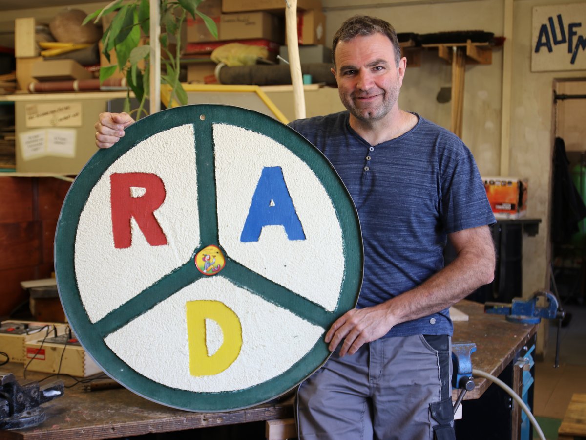 Lächelnder Mann in einer Werkstatt hält ein rundes Schild mit den Buchstaben R (rot), A (blau), D (gelb), die 'RAD' bilden.