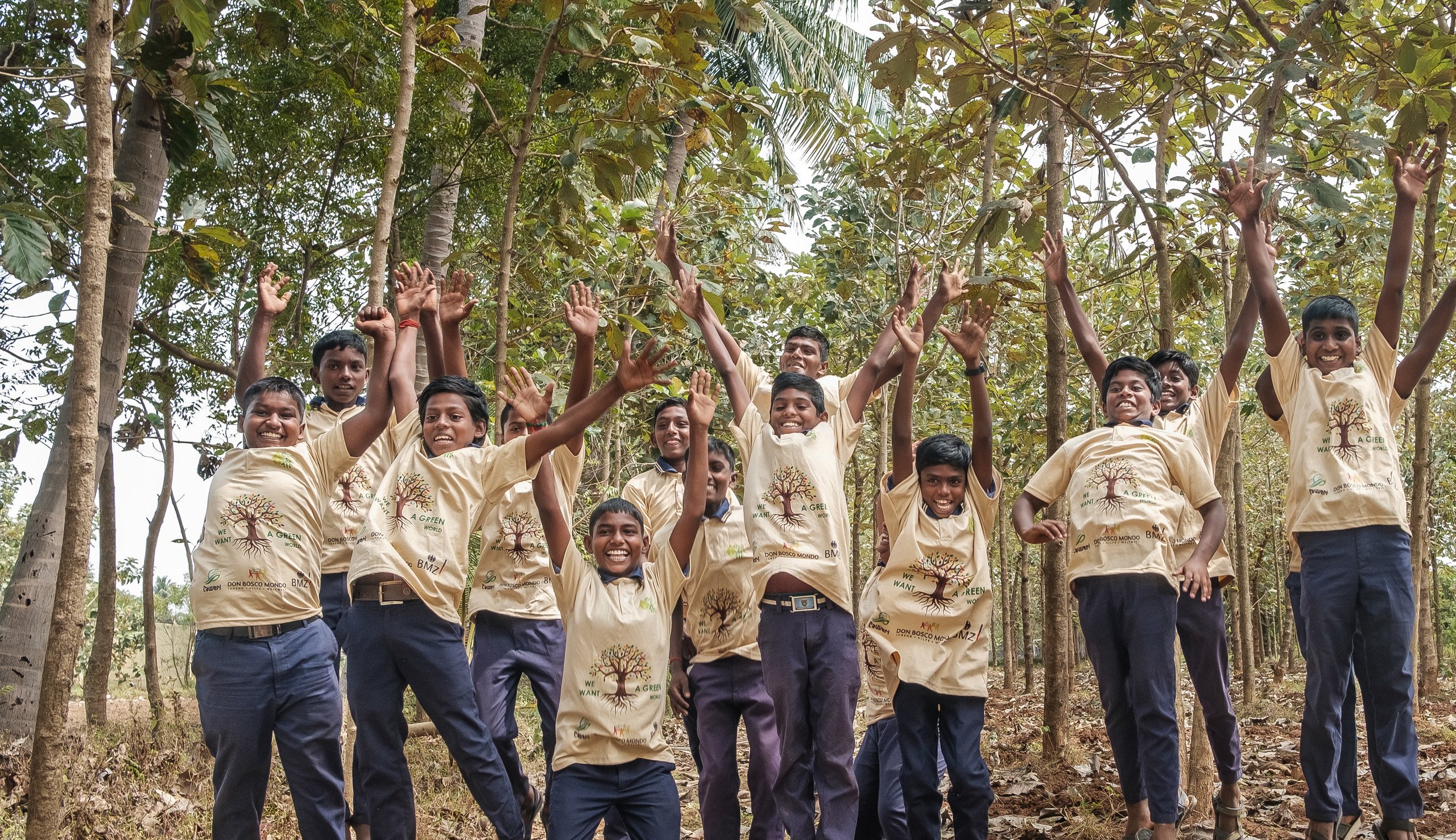 Eine Gruppe lachender Jungen springt in einem Wald und trägt T-Shirts mit dem Logo „WE WANT A GREEN WORLD“.