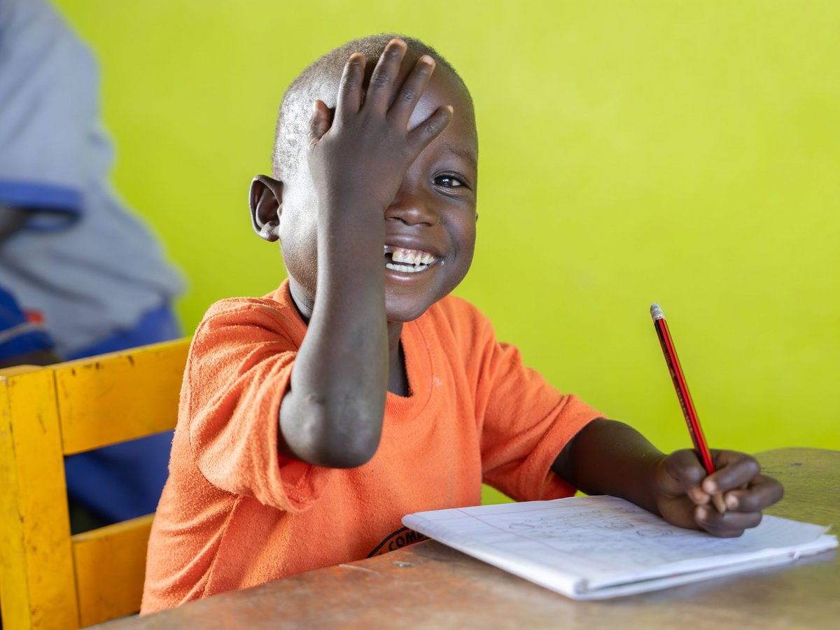 Ein lächelndes afrikanisches Kind im orangefarbenen Hemd bedeckt einen Teil des Gesichts mit einer Hand und hält einen Bleistift über einem Notizbuch.