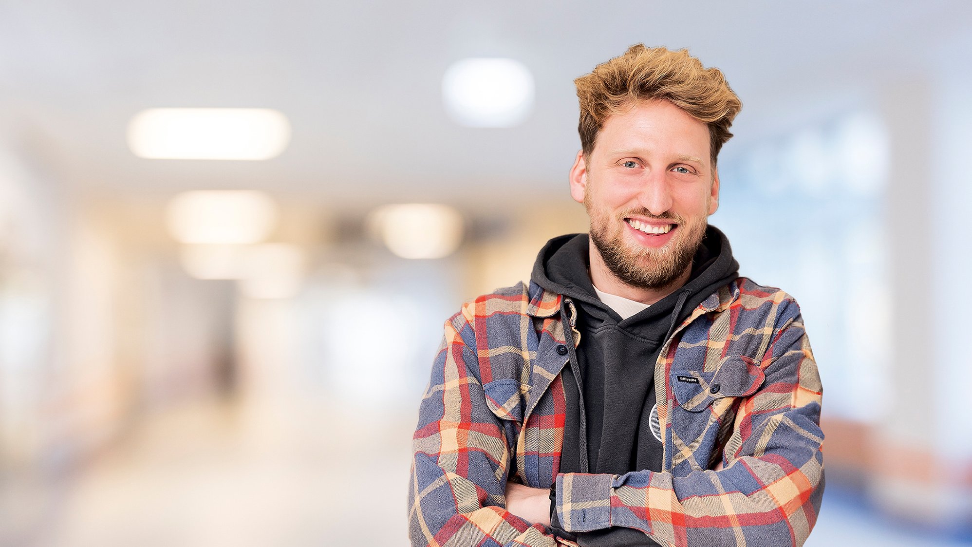 Lachender junger Mann mit Bart, trägt ein kariertes Hemd und Hoodie, Arme verschränkt vor unscharfem Innenhintergrund.