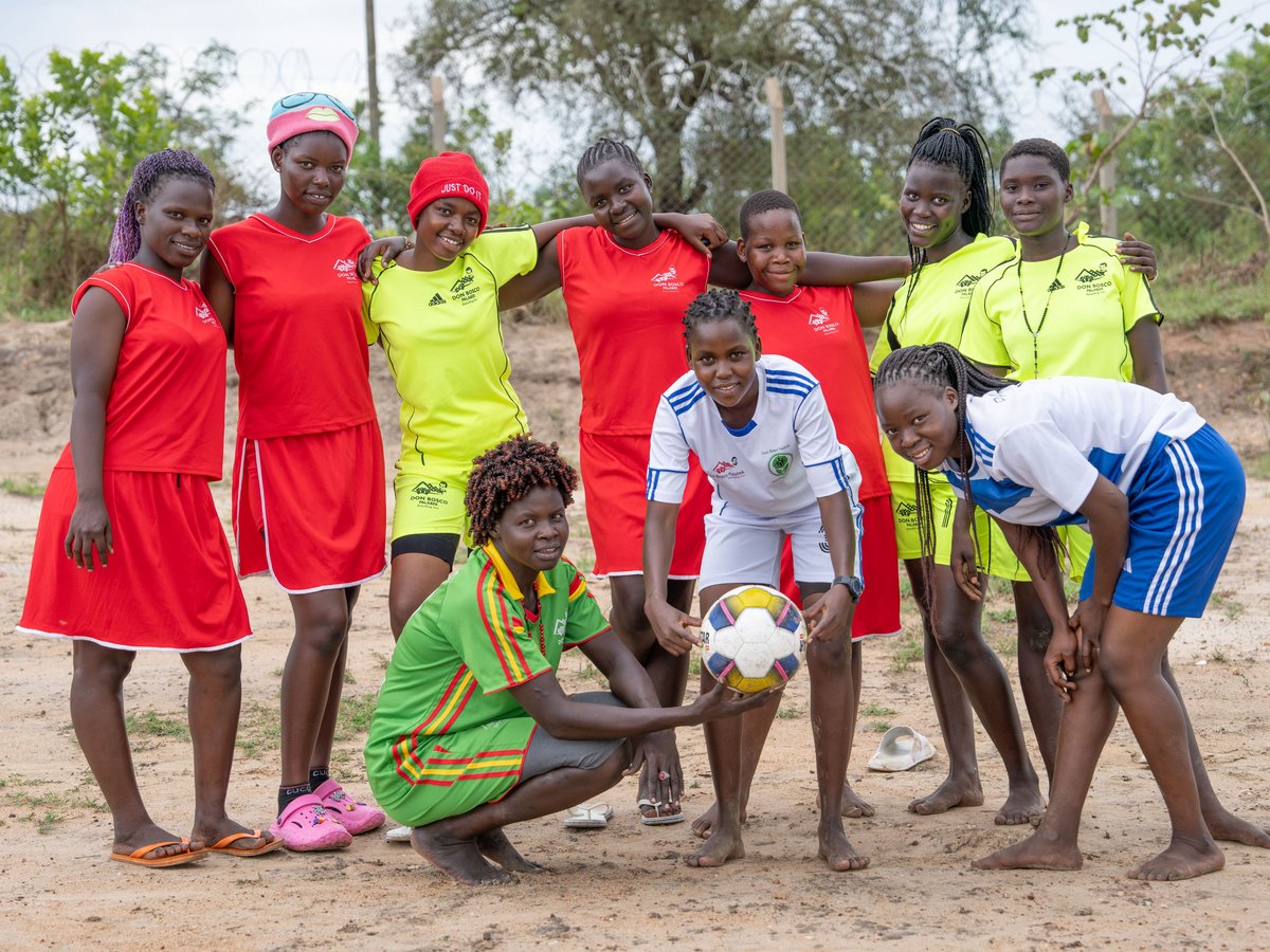Lächelnde Gruppe afrikanischer Mädchen in Sportkleidung, einige mit Fußball, posiert gemeinsam auf einem Sandplatz.