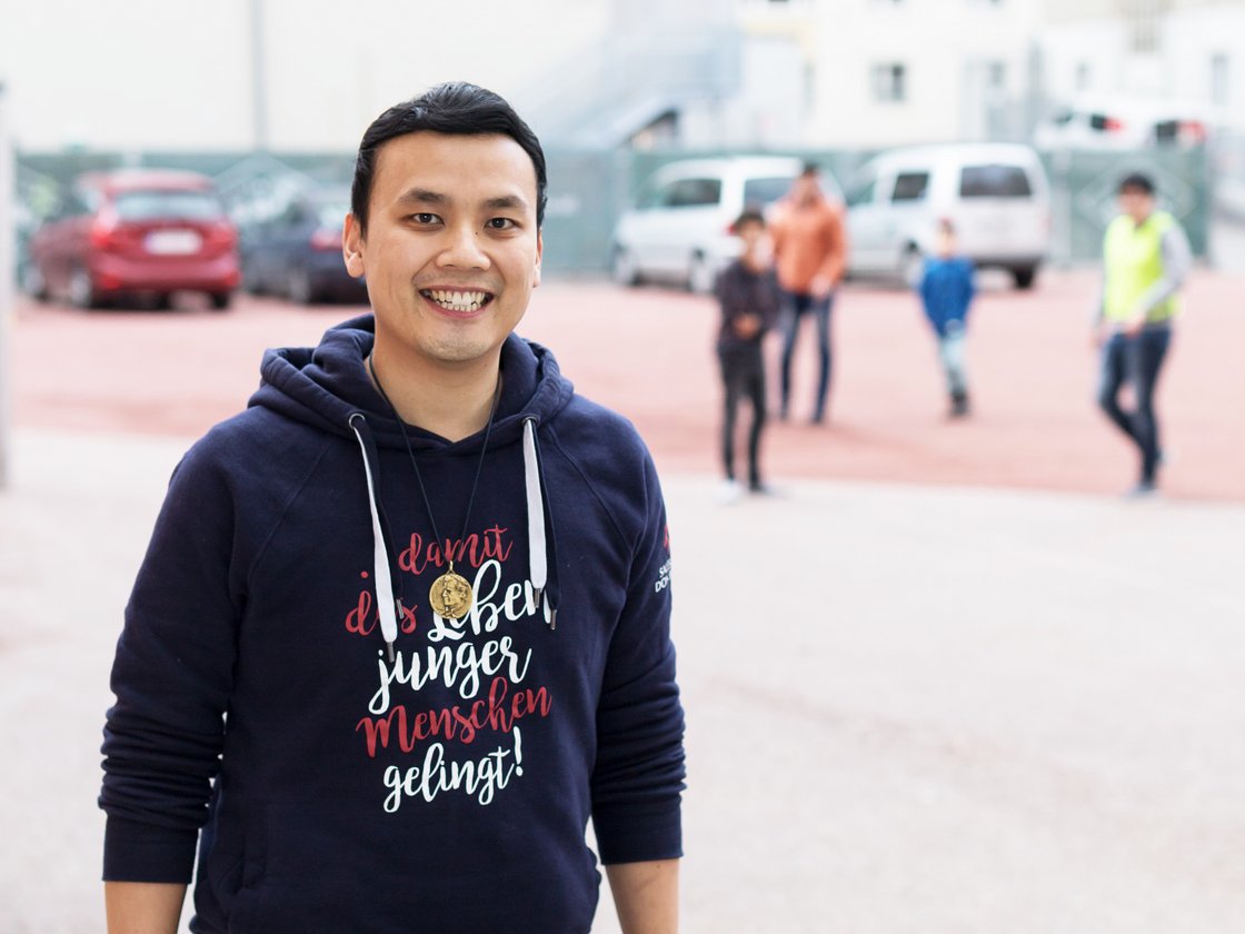 Lächelnder asiatischer Mann in blauem Hoodie mit deutschem Text 'damit das Leben junger Menschen gelingt!', im Freien.