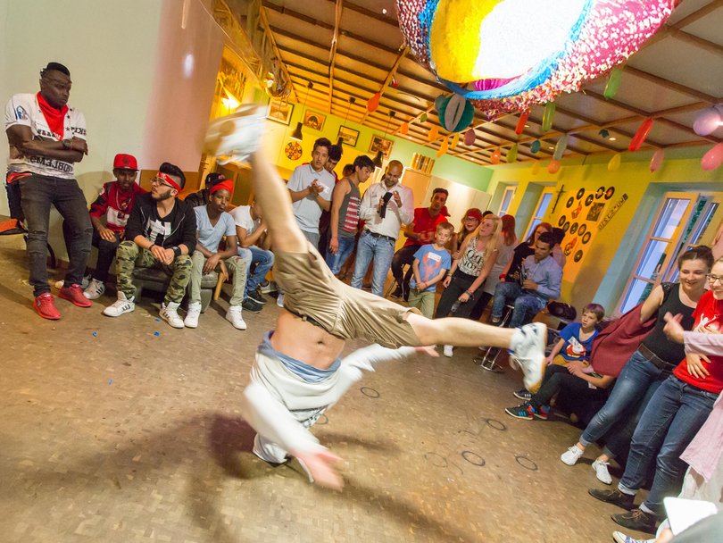 Ein Breakdancer führt einen akrobatischen Handstand-Move vor einer jubelnden Menge in einem festlich geschmückten Raum auf.