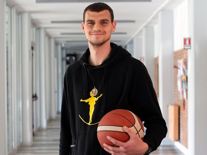 Lächelnder junger Mann in einem schwarzen Kapuzenpullover, der einen Basketball in einem modernen Flur hält.