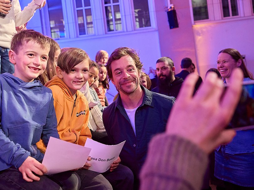 Chekcer Tobi sieht in eine Handykamera, daneben sitzen Kinder