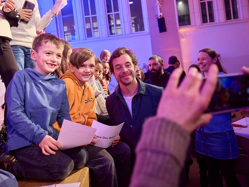 Chekcer Tobi sieht in eine Handykamera, daneben sitzen Kinder