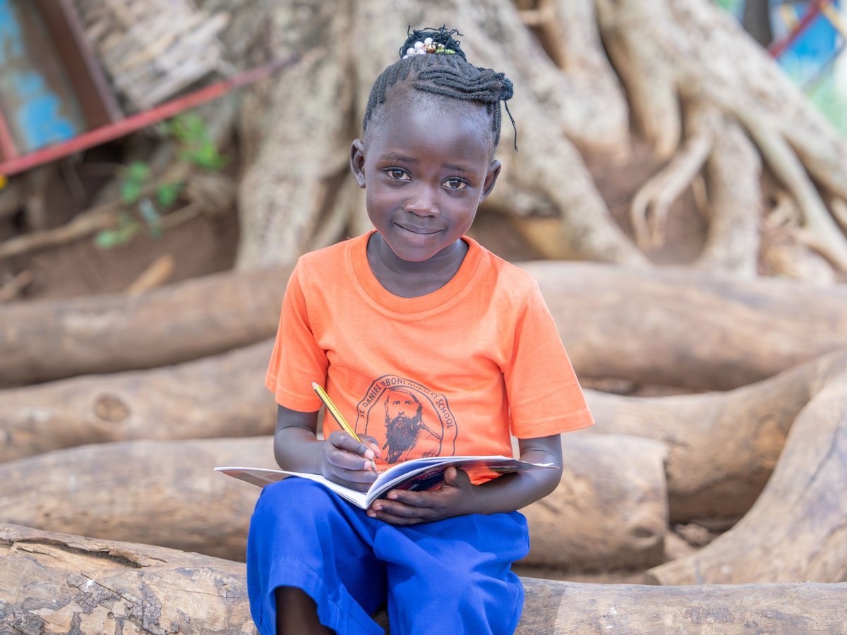 Lächelndes junges afrikanisches Mädchen sitzt draußen, schreibt in Notizbuch mit Bleistift. Trägt orangefarbenes Hemd und blaue Hose.