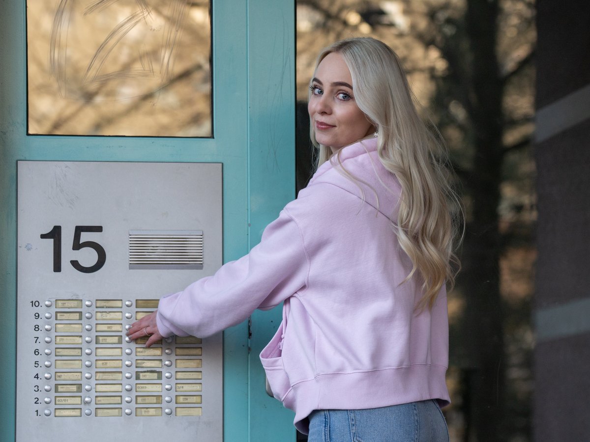 Blonde Frau in pinkfarbenem Hoodie drückt Klingelknopf an Gegensprechanlage mit Nr. 15, blickt zurück.