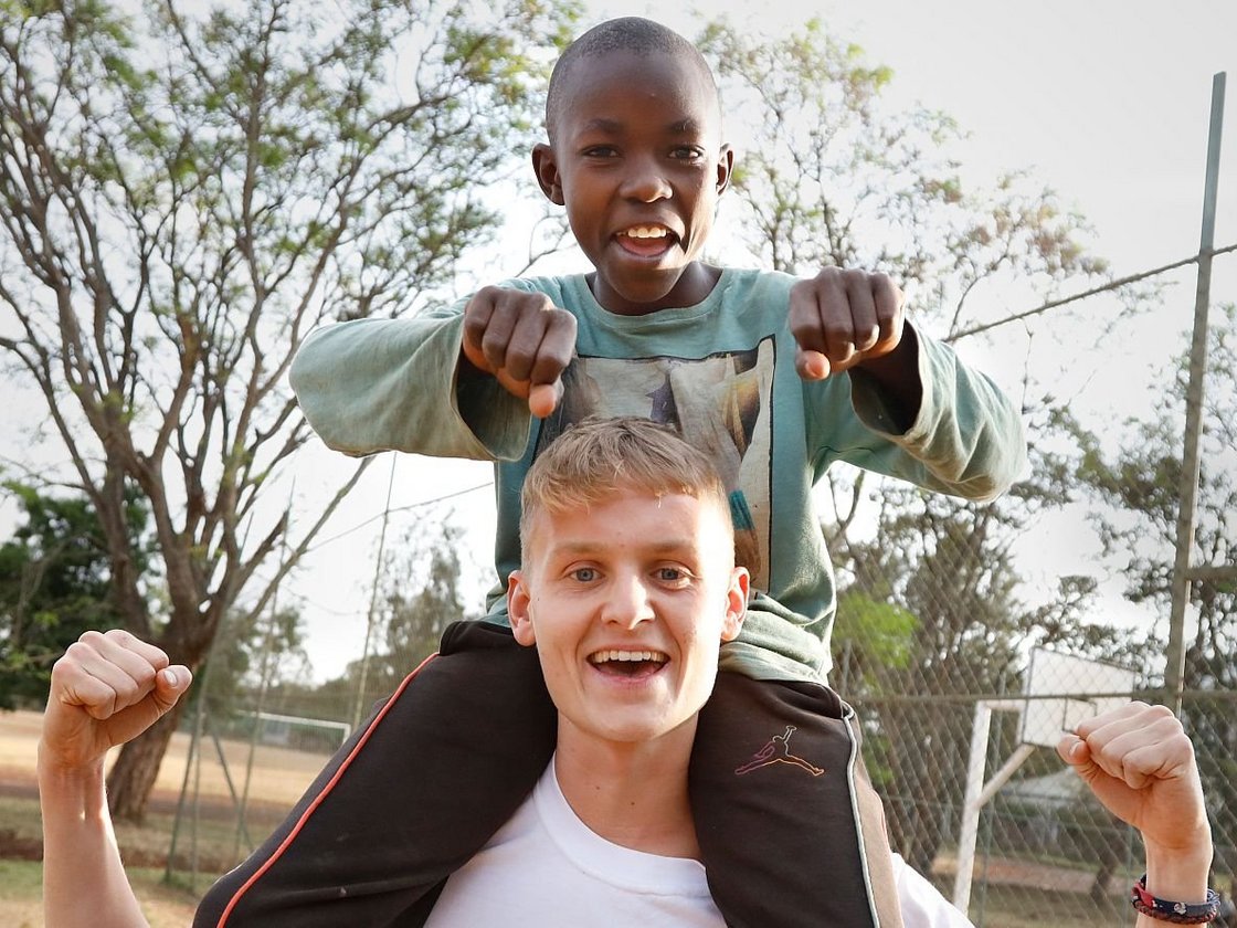 Ein lächelnder schwarzer Junge sitzt auf den Schultern eines fröhlichen weißen Mannes, beide heben spielerisch die Fäuste auf einem Sportplatz im Freien.
