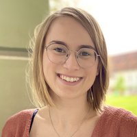Eine junge Frau mit schulterlangem blondem Haar und Brille lächelt strahlend in die Kamera, trägt einen rostroten Cardigan.