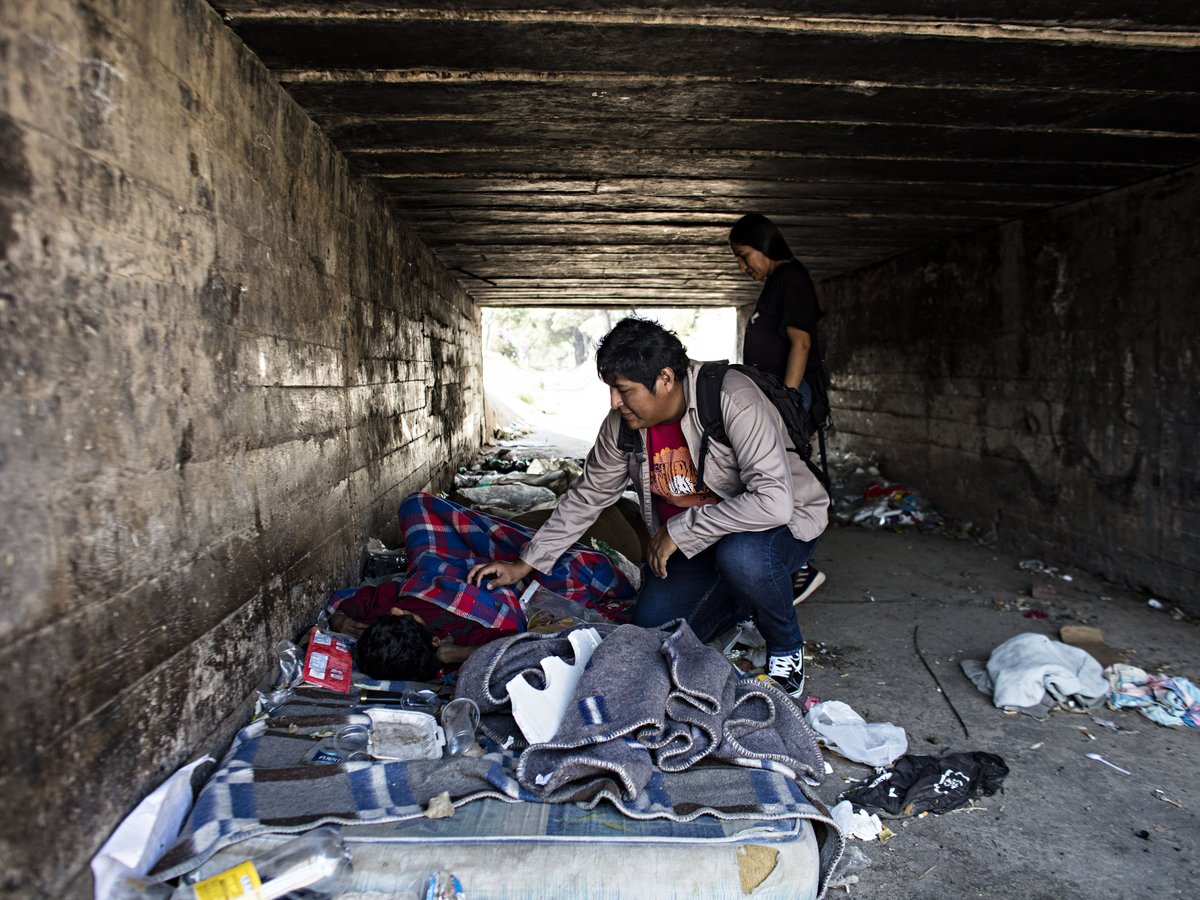 Drei Personen in einer Betonunterführung; eine liegt auf einem provisorischen Bett, eine kniet daneben, eine dritte steht.
