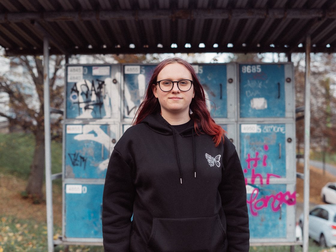 Junge Frau mit roten Haaren und Brille in schwarzem Hoodie vor einer mit Graffiti besprühten blauen Wand.