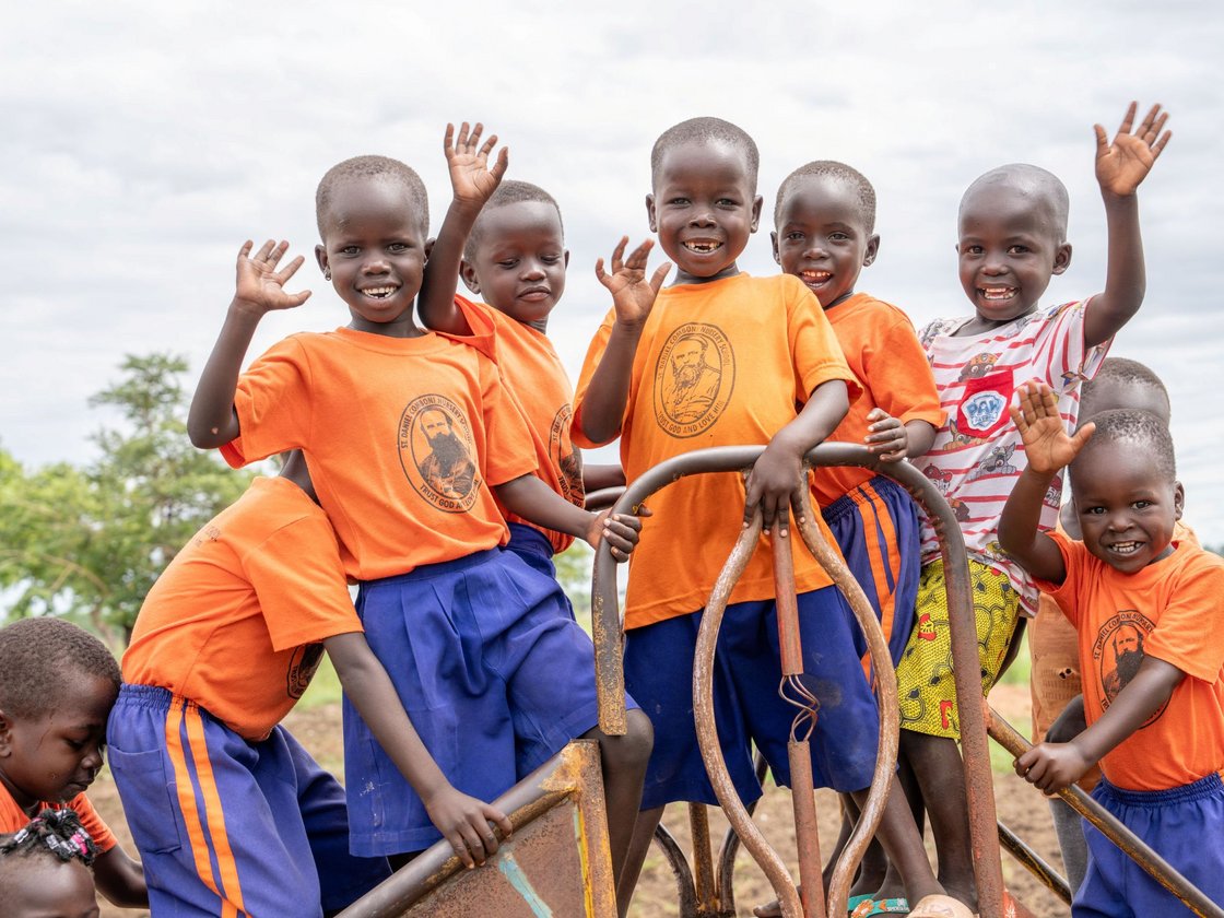 Fröhliche Kinder in orangefarbenen Schul-T-Shirts winken und lächeln auf einem Spielplatz.