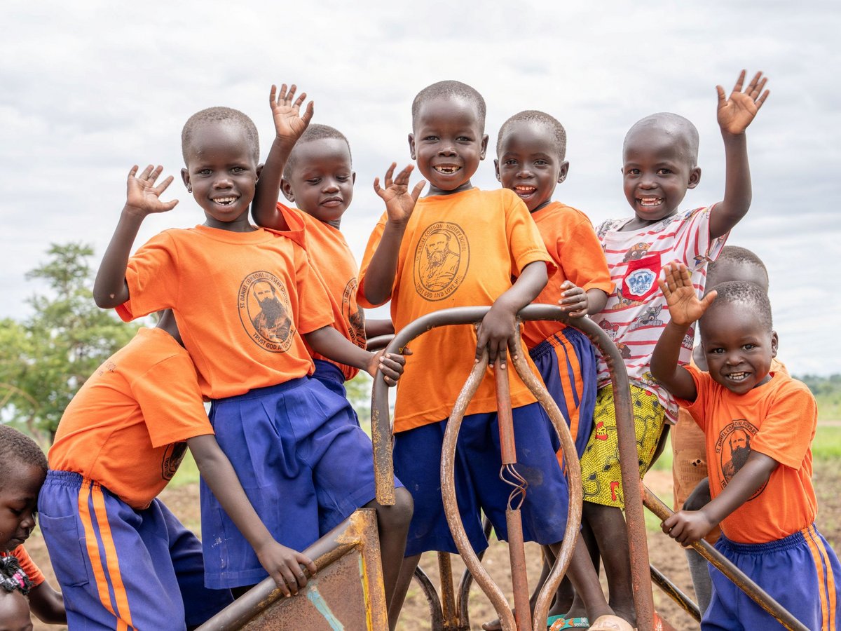 Fröhliche Kinder in orangefarbenen Schul-T-Shirts winken und lächeln auf einem Spielplatz.