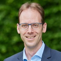 Porträt eines lächelnden Mannes mit braunen Haaren und Brille, hellblauem Hemd und dunklem Sakko, vor grünem Hintergrund.