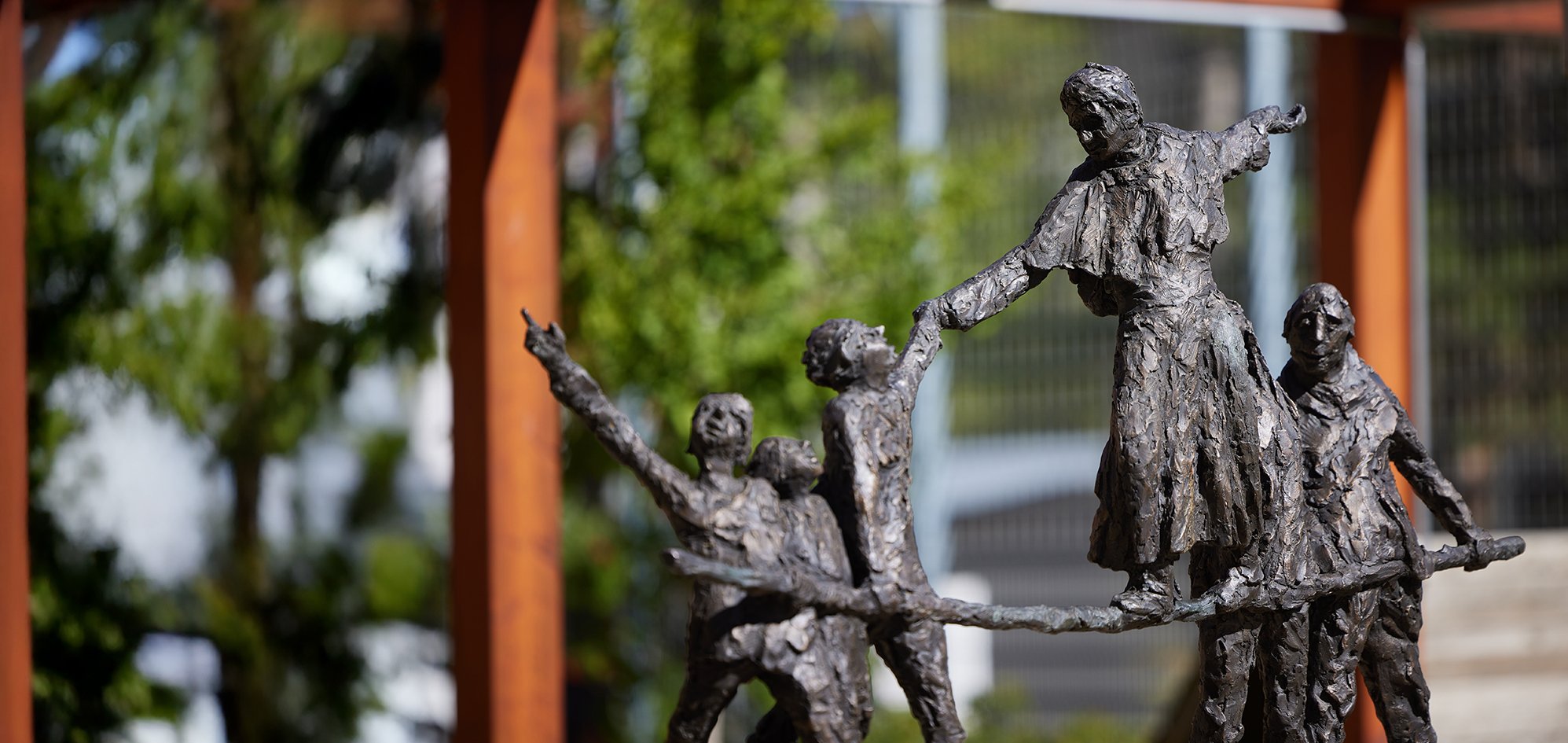 Texturierte Bronzeskulptur mehrerer Figuren, einige zeigen und reichen, vor verschwommenem Außenhintergrund.