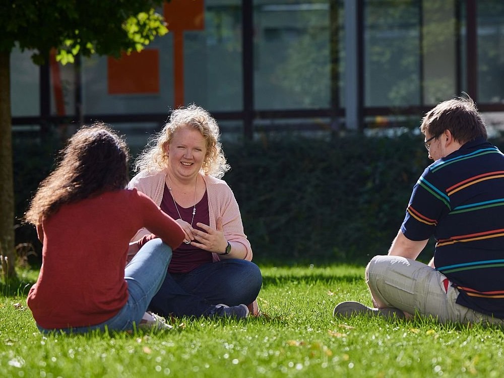 Drei Personen sitzen an einem sonnigen Tag draußen auf einer Wiese, eine Frau lächelt eine andere während eines Gesprächs an.