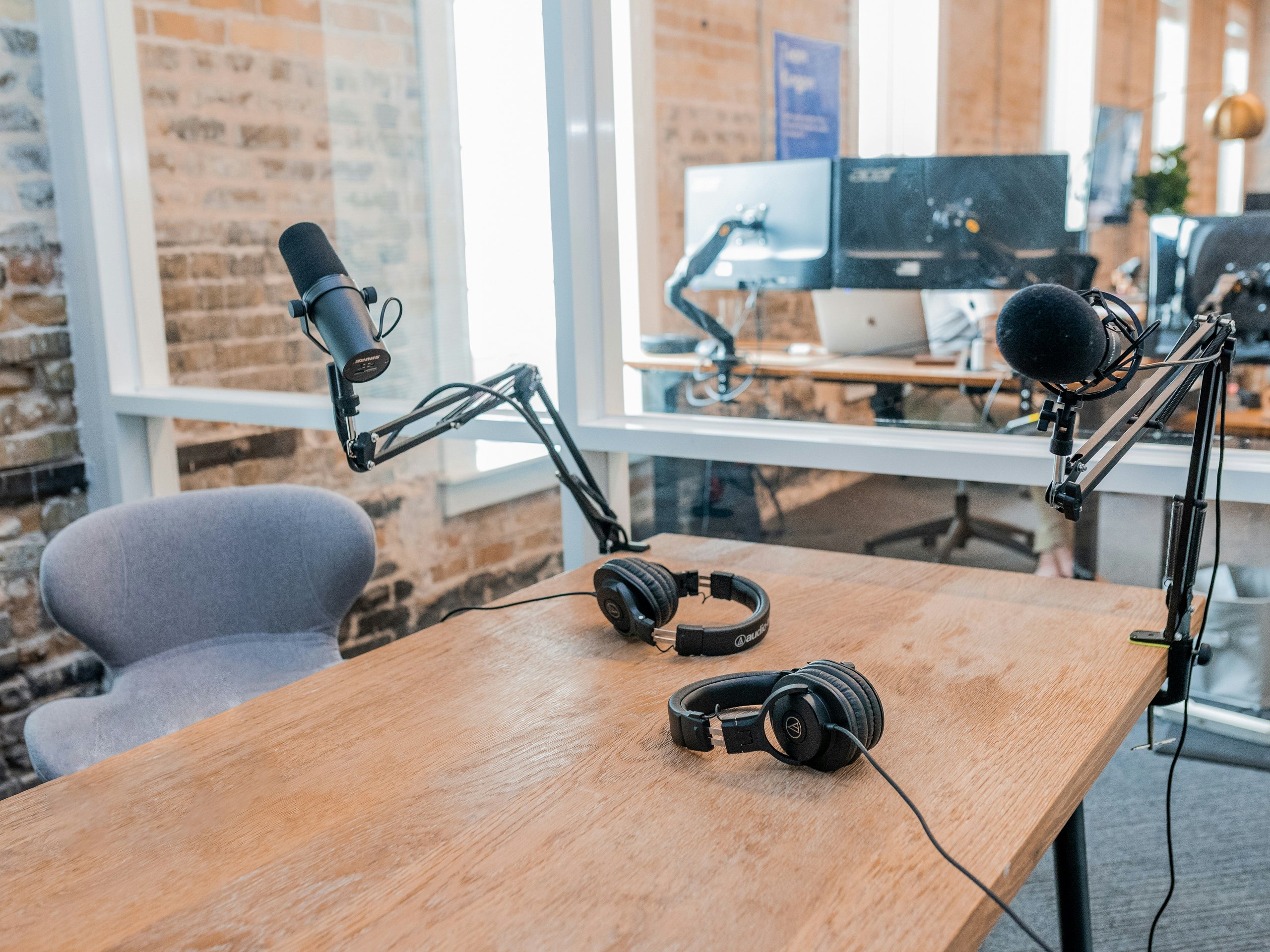 Podcast-Studio-Setup mit zwei Mikrofonen, zwei Kopfhörern und einem grauen Stuhl an einem Holztisch im Büro.