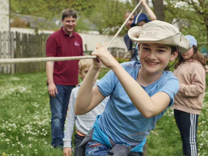 Lächelnder Junge mit Hut hält ein Seil bei einer Outdoor-Gruppenaktivität mit anderen Kindern und einem Erwachsenen.