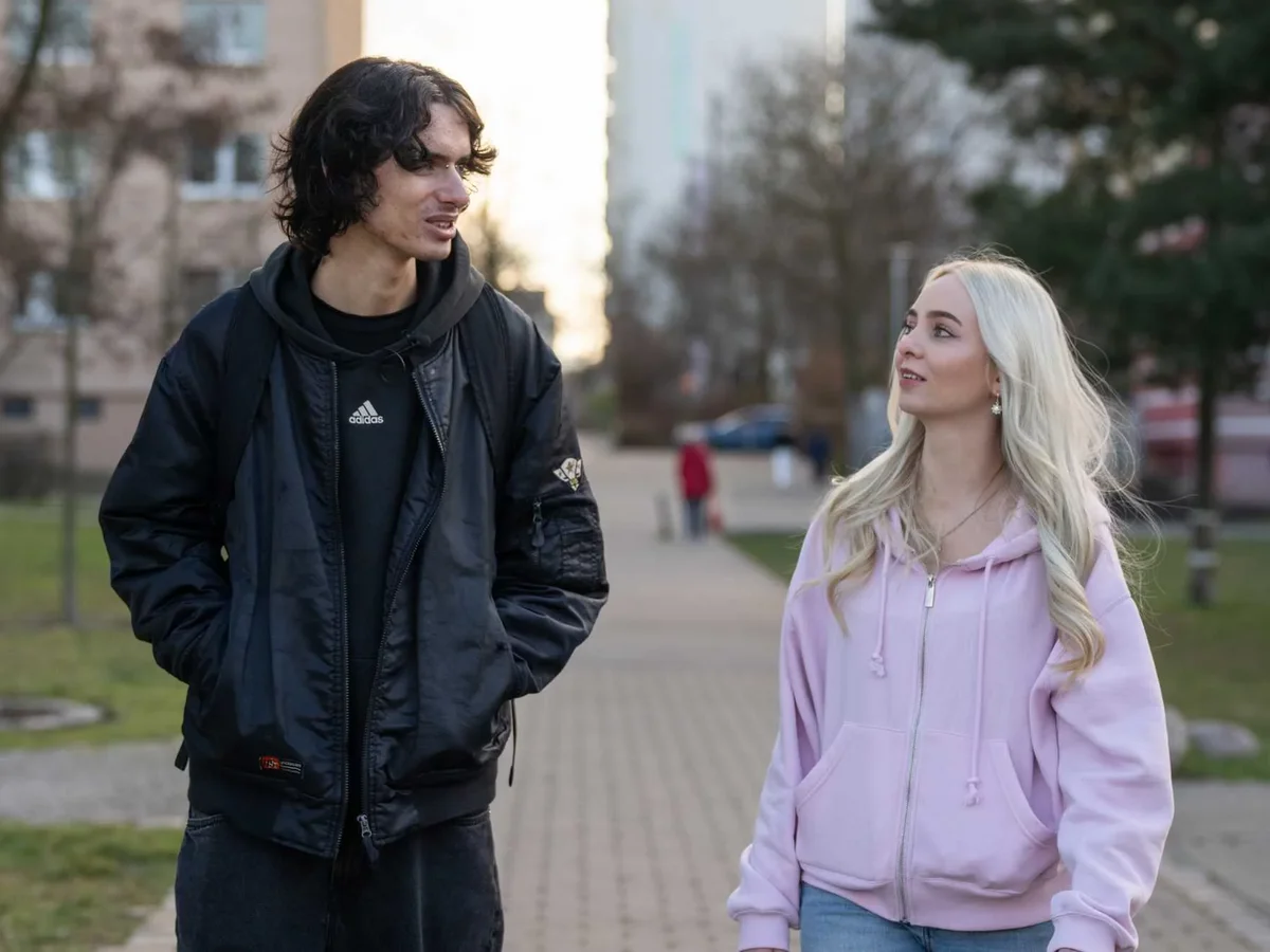Ein Mann mit dunklen Haaren und eine blonde Frau in rosa Hoodie spazieren im Freien und unterhalten sich.