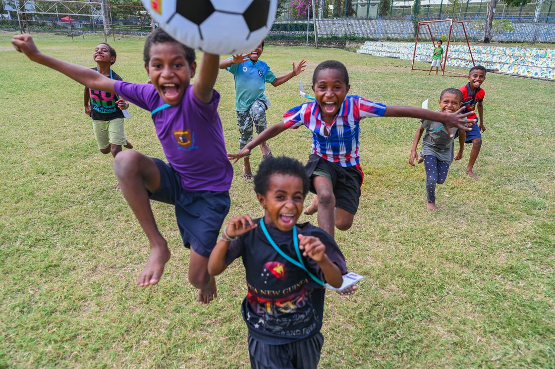 Fröhliche Kinder spielen Fußball auf einem Rasenplatz. Ein Kind springt mit dem Ball über dem Kopf, umgeben von anderen begeisterten Kindern.