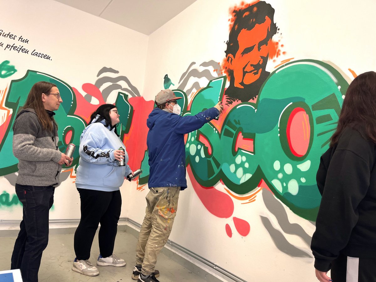 Gruppe von Personen malt gemeinsam ein farbenfrohes Graffiti-Wandbild auf einer weißen Wand, mit Porträt und abstrakten Formen.