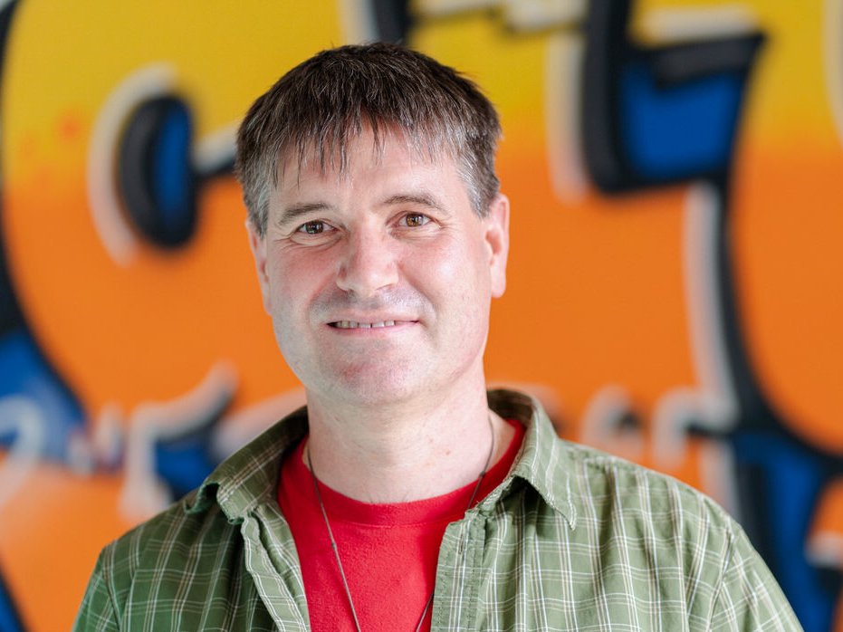 Ein lächelnder Mann mit kurzen braunen Haaren und einem grünen Karoshirt über einem roten T-Shirt vor einer unscharfen Graffiti-Wand.