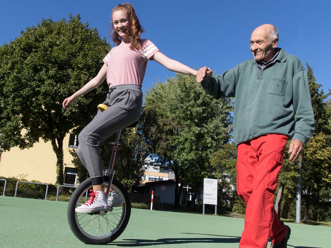 Eine junge Frau auf einem Einrad hält Händchen mit einem älteren Mann, der ihr lächelnd auf einem grünen Platz hilft.