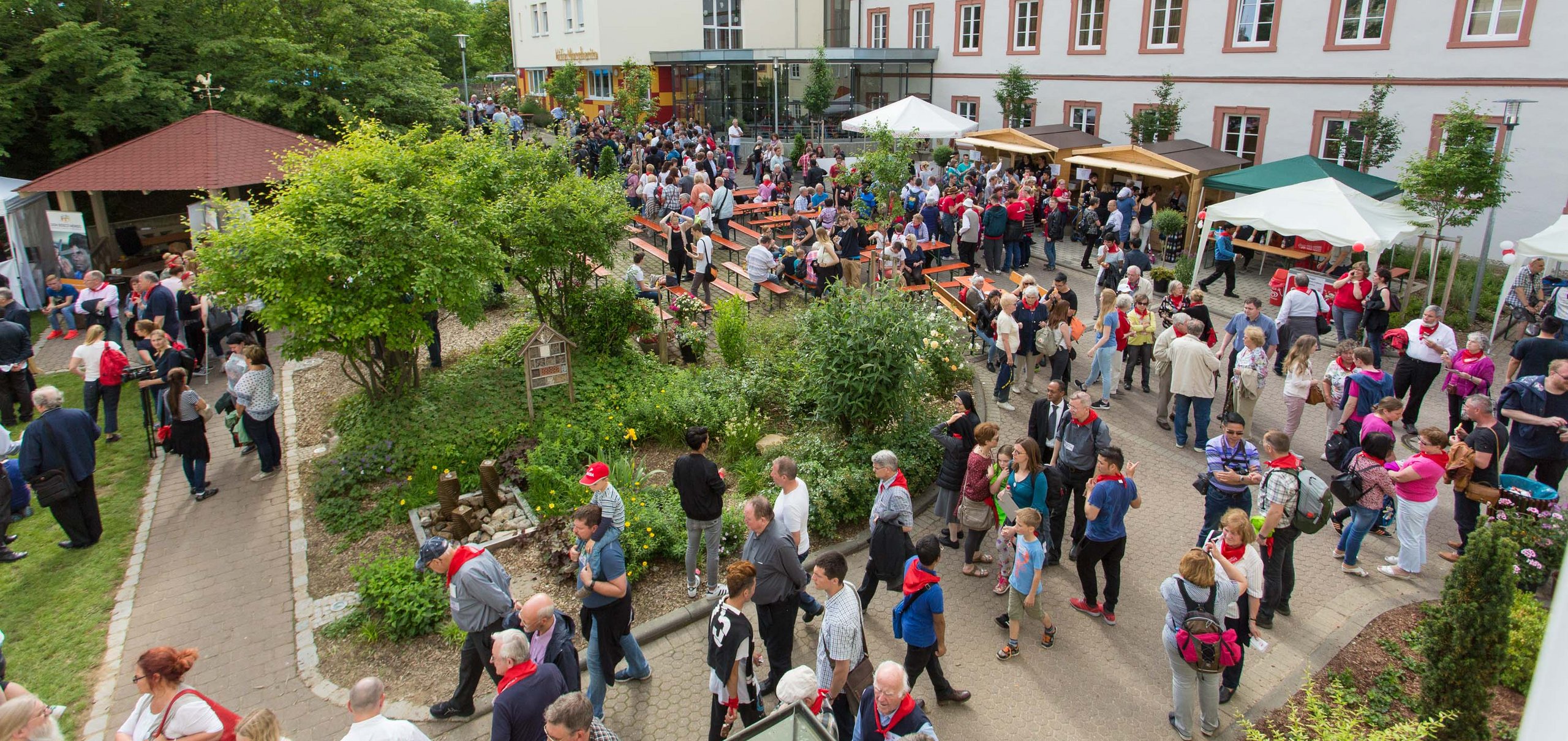 Viele Menschen bei einer lebhaften Outdoor-Veranstaltung in einem grünen Gartenhof mit Gebäuden, Bänken und Ständen.