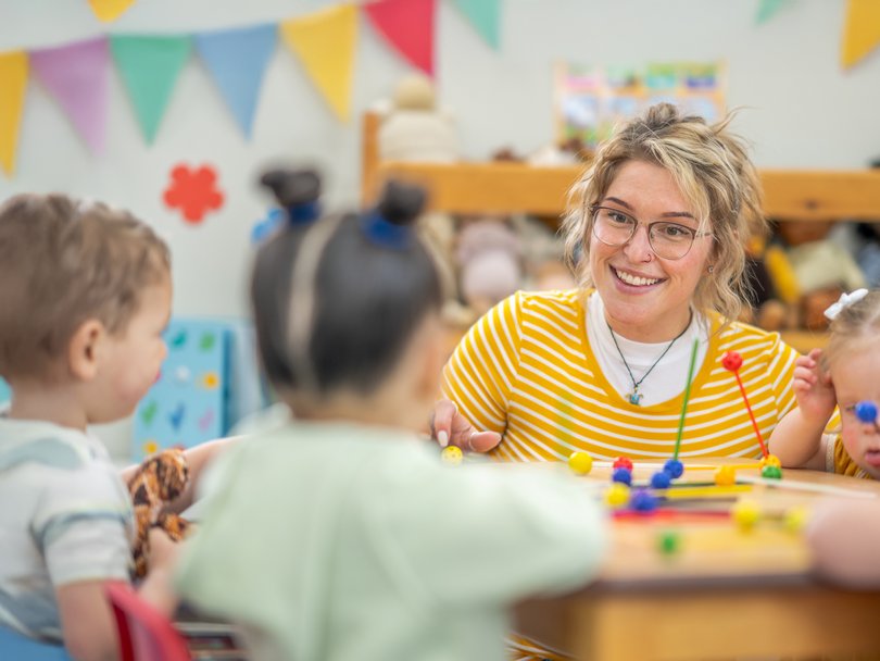 Eine lächelnde Erzieherin mit Brille und blonden Haaren sitzt mit Kleinkindern an einem Tisch, die mit bunten Spielsachen spielen.