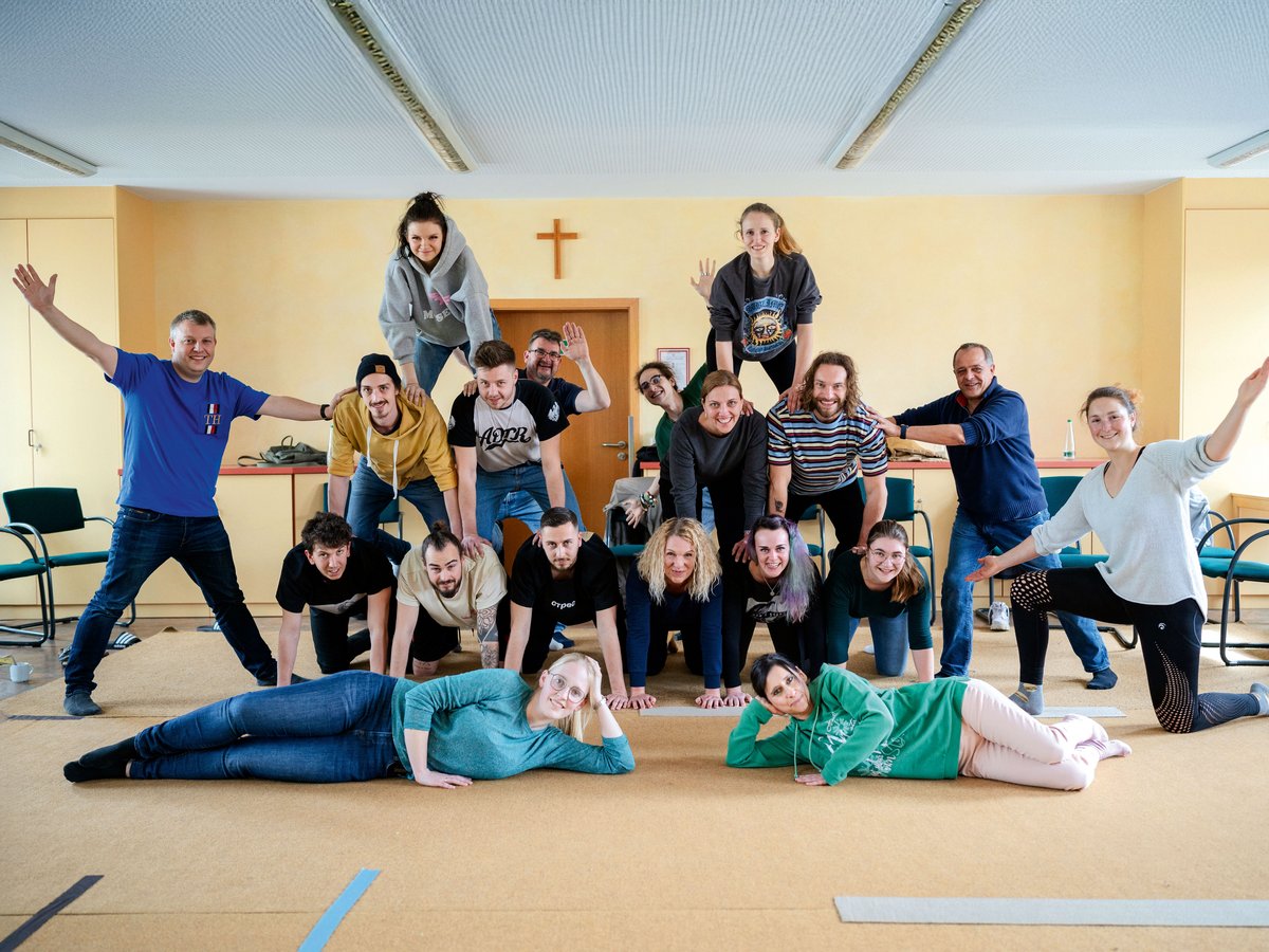 Eine große Gruppe lächelnder Menschen bildet eine Menschenpyramide in einem hellen Raum.