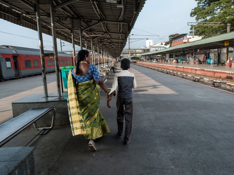 Eine indische Frau im Sari hält die Hand eines Jungen, sie gehen über einen Bahnsteig mit einem wartenden Zug.