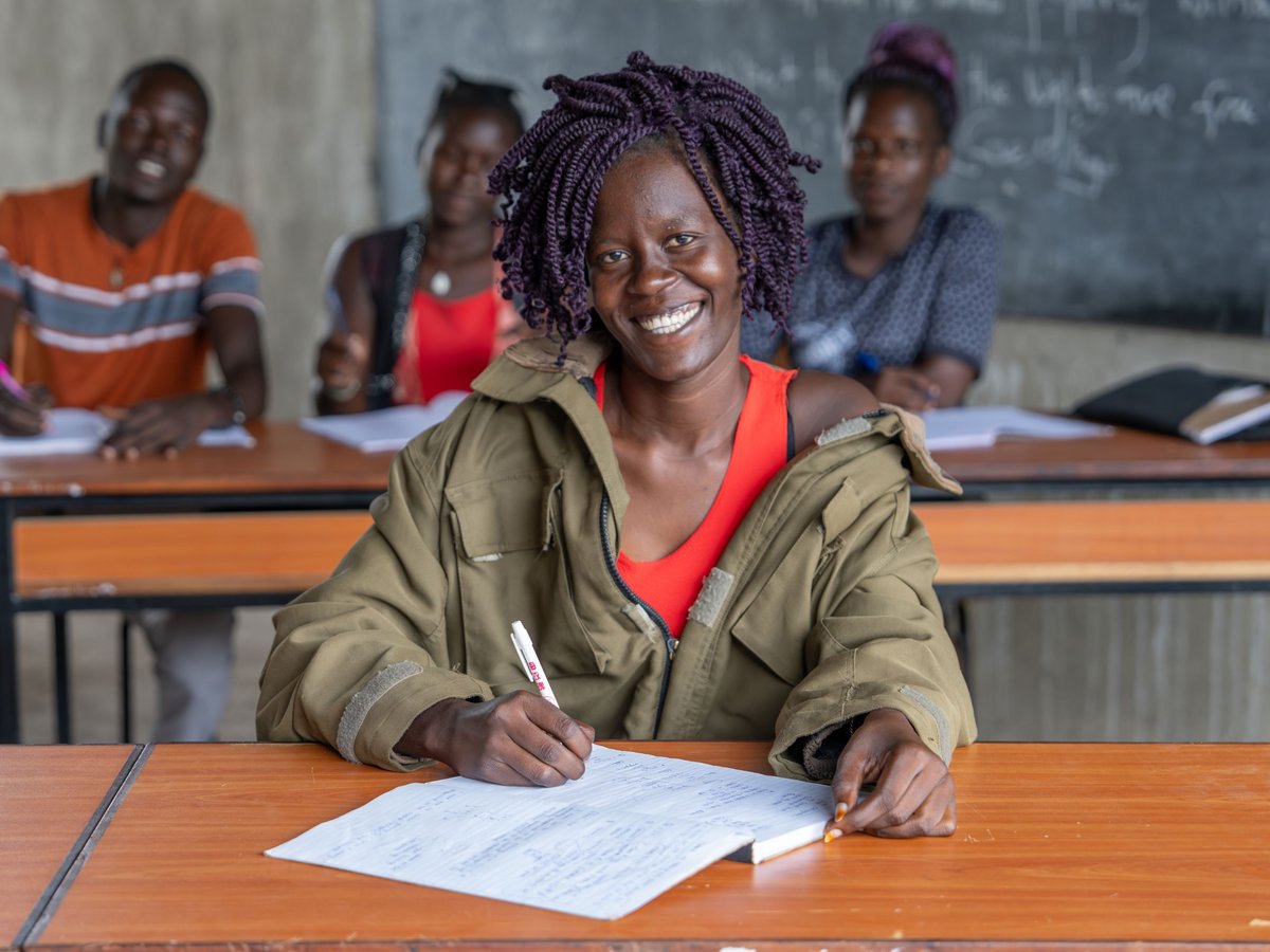 Eine lächelnde junge Frau mit lila Dreadlocks schreibt in einem Klassenzimmer, andere Schüler und eine Tafel im Hintergrund.
