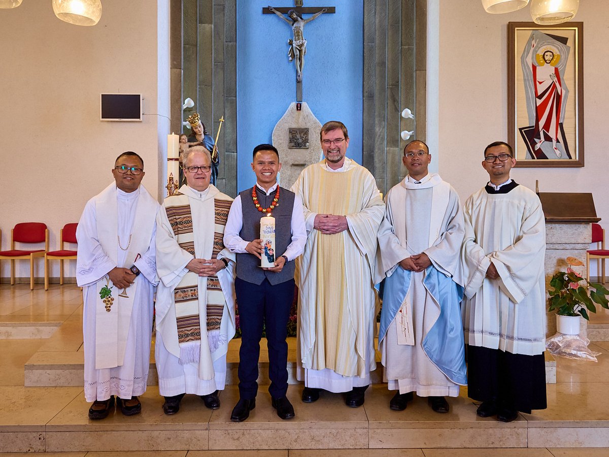 Sechs lächelnde Männer in geistlichen Gewändern posieren in einer Kirche. Einer hält eine verzierte Kerze. Kruzifix und Kunst im Hintergrund.