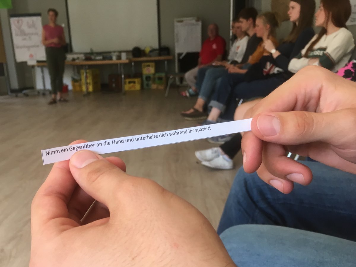 Hände halten Papierstreifen mit der Anweisung: "Nimm ein Gegenüber an die Hand und unterhalte dich während ihr spaziert".