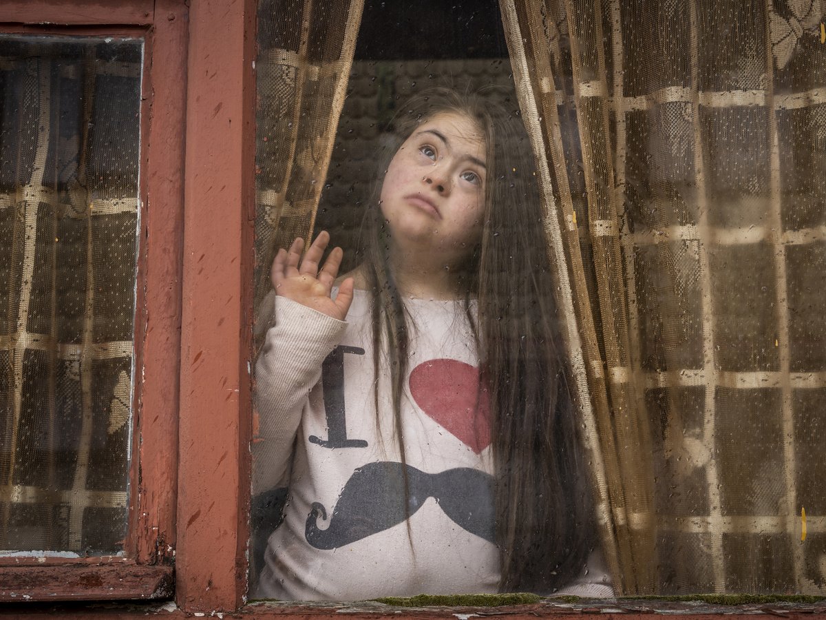 Ein Mädchen mit langen Haaren blickt durch ein regennasses Fenster, Hand am Glas, trägt Herz-Schnurrbart-Shirt.